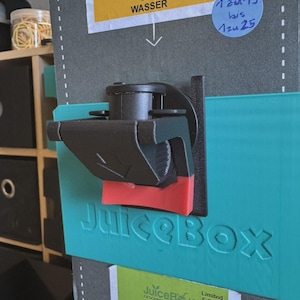 Peut inclure: Un distributeur de boissons noir et rouge fixé à un panneau turquoise avec le mot "JuiceBox" en relief. L'image comprend des étiquettes avec le texte "SKI-WASSER" et "TROPICAL LOVE".