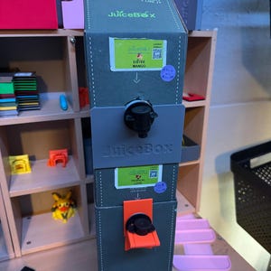 May include: A tall, rectangular, olive green JuiceBox dispenser featuring two stacked beverage taps. The top tap is black, and the bottom tap is orange. Each section has a label for Eistee Mango and Tropical Love, with JuiceBox branding.