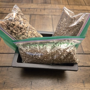 May include: Three clear plastic bags filled with different types of soil and potting mix, placed inside a dark gray rectangular container. The bags have green and purple zipper closures. The container sits on a wooden surface.