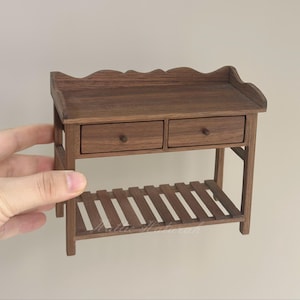 May include: A miniature wooden table with two drawers and a slatted shelf. The table is a rich brown color and has a scalloped edge along the top. The drawers have small, round knobs.