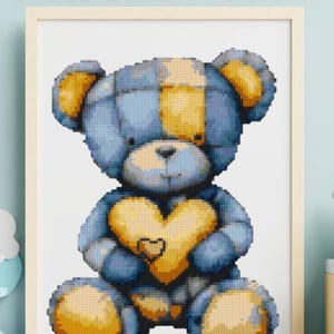 Op de afbeelding: Een ingelijst kruissteekpatroon met een blauw en gele teddybeer die een hart vasthoudt. De tekst "Cute Teddy Cross Stitch Pattern" staat bovenaan. Extra tekst is "Beginner Friendly PDF Download Easy Stitch".
