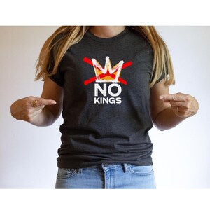 May include: Dark gray t-shirt with the text "NO KINGS" in white block letters. A red line crosses out a gold and white crown above the text. The shirt is worn with blue jeans.