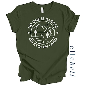 May include: An olive green t-shirt with the text "NO ONE IS ILLEGAL ON STOLEN LAND" in a white circular design featuring mountains, trees, and a river. The shirt is made of a soft material.