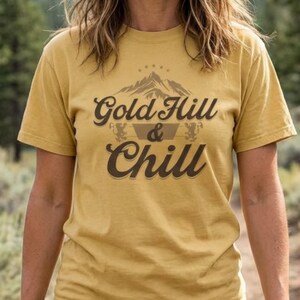 May include: Mustard-coloured t-shirt featuring the text "Gold Hill & Chill" and a mountain graphic. The shirt is worn by a person in blue jeans, walking on a path in a natural setting. A mockup of the shirt is also displayed.