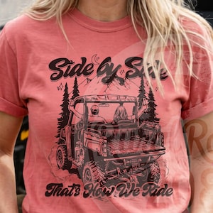 Könnte beinhalten: Wassermelonfarbenes T-Shirt mit einer Grafik eines Side-by-Side-Fahrzeugs und dem Text "Side by Side, That's How We Ride." Das Shirt wird vor einem Side-by-Side-Fahrzeug gezeigt.