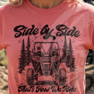Könnte beinhalten: Wasserfarbenes T-Shirt mit einem schwarzen Aufdruck eines Side-by-Side-Fahrzeugs, Bäumen und dem Text "Side by Side" und "That's How We Ride." Ein kleineres Mockup des Shirts ist ebenfalls sichtbar. Das Shirt hat einen Rundhalsausschnitt.