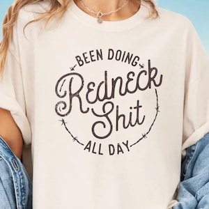 Könnte beinhalten: Elfenbeinfarbenes T-Shirt mit dem Text "Been Doing Redneck Shit All Day" in einem kreisförmigen Design. Der Text ist in einer Used-Schriftart gehalten. Das T-Shirt ist kurzärmelig. Ein Mockup ist ebenfalls zu sehen.