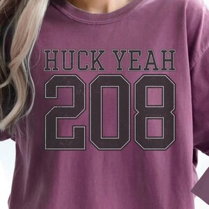 Op de afbeelding: Bessenkleurig T-shirt met de tekst "HUCK YEAH 208" in een zwarte, collegiale lettertype. Een kleinere mockup van het shirt is rechtsonder zichtbaar. Het shirt heeft een relaxte pasvorm.