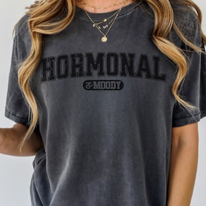 Può includere: T-shirt color grafite con la scritta "HORMONAL & MOODY" in nero, con un carattere in stile collegiale. La maglietta è di colore grigio scuro e ha un aspetto vintage e usurato. Una tazza di plastica trasparente con cannuccia e bevanda marrone.