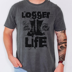 以下が含まれることがあります： 色あせたグラフィックのダークグレーのTシャツ。デザインは、作業ブーツのペアの上に「LOGGER」という単語、両側に「DIRT BOOTS」と「STEAM MONEY」が特徴です。「LIFE」という単語がブーツの下に印刷されています。