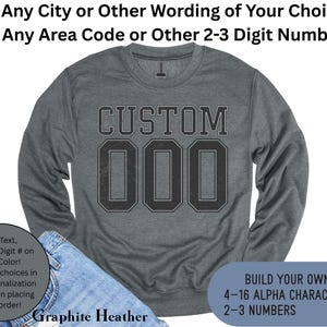 May include: Graphite heather sweatshirt with the word "CUSTOM" and the numbers "000" in a black, collegiate-style font. The sweatshirt has long sleeves and a crew neck. Text on the image says "Build Your Own!" and "Any Text, Any 2-3 Digit # on Any Color!"