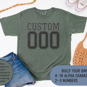 May include: A moss green t-shirt with the word "CUSTOM" and the numbers "000" printed in black. The shirt is displayed with denim shorts, a woven bag, and white sneakers. Text on the image says "Build Your Own!" with character and number specifications.