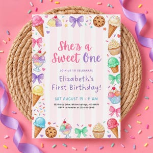 May include: A pink and white invitation card with the text "She's a Sweet One" and "Elizabeth's First Birthday!" surrounded by ice cream cones, cupcakes, and bows. The card is set on a woven mat with pink sprinkles and a purple ribbon.
