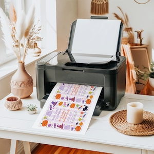 May include: A black printer printing a Halloween party invitation. The invitation has a purple background with orange pumpkins, purple witch hats, and ghosts. The text on the invitation reads "Too Cute to Spook". The printer is on a white table.