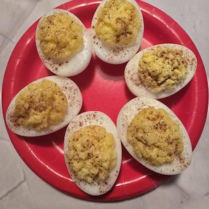 May include: Six deviled eggs arranged on a red paper plate. The eggs are cut in half, with a yellow filling and a sprinkle of dark seasoning. The white egg whites are visible.