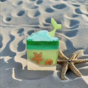 May include: A green, blue, and white layered soap bar with a mermaid tail on top. The soap bar is decorated with a seashell and a starfish. The soap bar is sitting on a sandy surface.