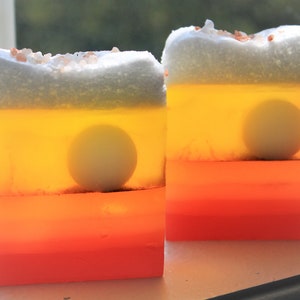 May include: Two handmade soap bars with a layered design. The top layer is white with a light pink hue, the second layer is yellow, the third layer is orange, and the bottom layer is a darker pink. Each bar has a white circle embedded in the yellow layer.