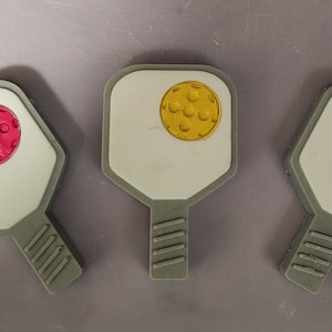 May include: Three white pickleball paddles with different colored pickleballs on them. The paddles are gray and have a textured grip. The pickleballs are red, yellow, and green.