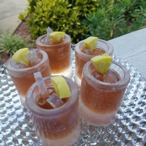 May include: Five clear glass shot glasses filled with a brown liquid and ice cubes. Each glass has a yellow lemon wedge on top.