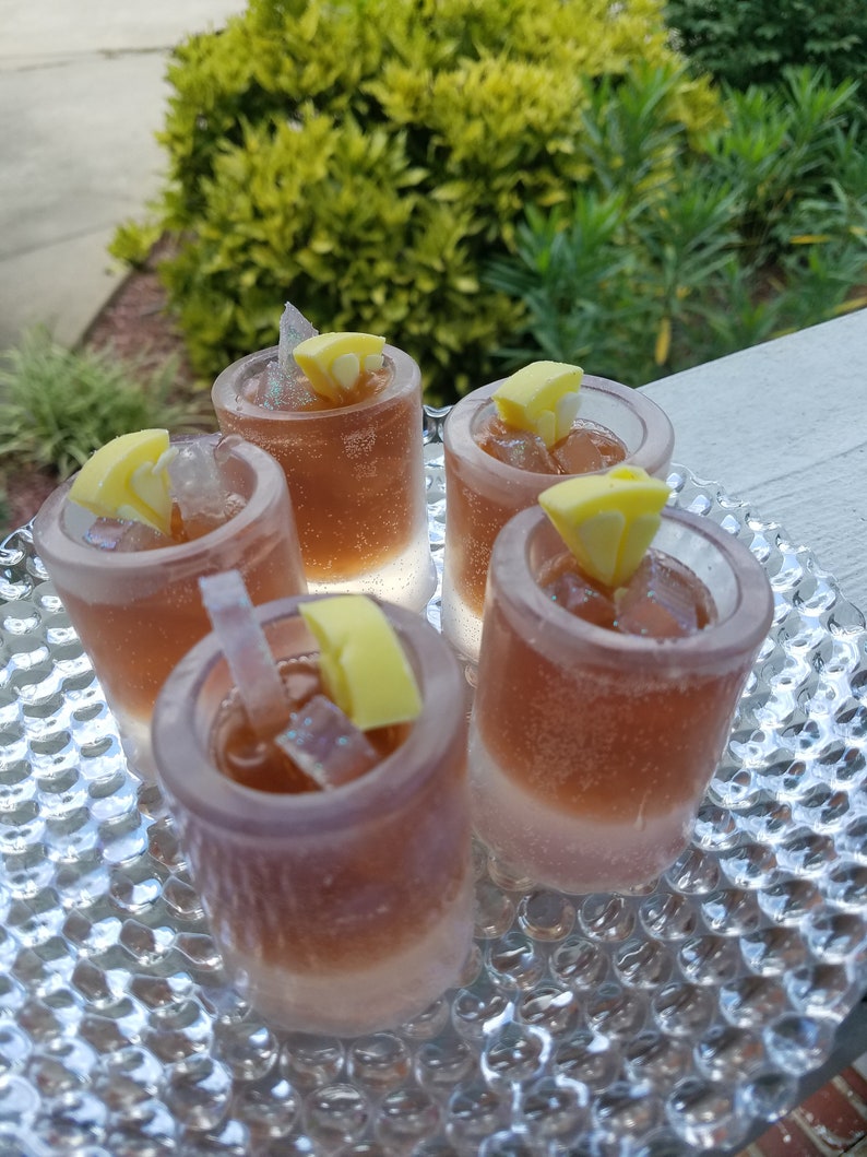 May include: Five clear glass shot glasses filled with a brown liquid and ice cubes. Each glass has a yellow lemon wedge on top.