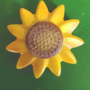 May include: A sunflower-shaped object featuring bright yellow, smooth petals radiating from a central, textured, glittery brown disc. The design is symmetrical, with a distinct contrast between the vibrant yellow petals and the dark, sparkling center.