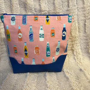 May include: A pink zippered pouch with a blue bottom. The pouch is decorated with a pattern of colorful soda bottles with various labels, including "FIZZLE", "SODA", "SPORT", "FRESH", and "7UP".