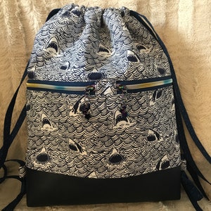 May include: A black and white drawstring backpack with a shark and wave pattern. The backpack has a black bottom and two zippered pockets on the front.