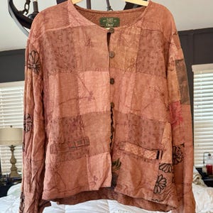 May include: A long-sleeved button-down shirt in shades of dusty rose and terracotta, featuring a patchwork design with varied textures and subtle floral embroidery. The garment has a round neckline, a front button closure, and two front pockets.