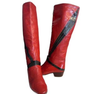 May include: A pair of knee-high boots in bright red with a smooth finish. Each boot features a dark, textured snakeskin-patterned accent strip that runs diagonally across the shaft and wraps around the ankle area. The boots have a low block heel.