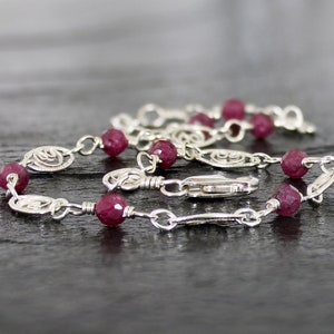 May include: A silver chain bracelet with small, round, dark pink gemstones. The chain features a delicate, ornate design.