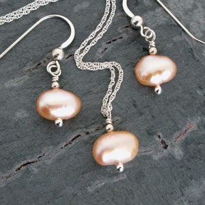 May include: A delicate silver necklace and matching earrings feature a pair of soft pink oval pearls. The necklace has a delicate chain and the earrings have simple hook closures.