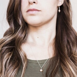 May include: A woman with long brown hair wearing a green shirt and a silver necklace with a small pearl pendant. She is also wearing small silver earrings with a pearl accent.