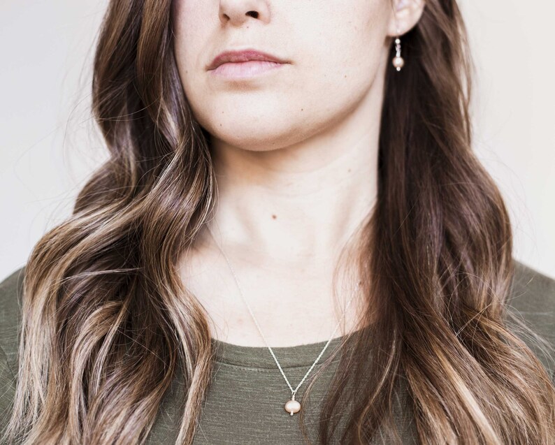 May include: A woman with long brown hair wearing a green shirt and a silver necklace with a small pearl pendant. She is also wearing small silver earrings with a pearl accent.