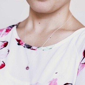 Puede incluir: Un collar de cadena de plata con un peque&ntilde;o colgante redondo de piedra rosa. El collar lo lleva una persona que lleva una blusa blanca con estampado floral.