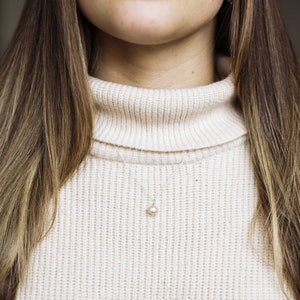 May include: A close-up of a woman wearing a cream-colored turtleneck sweater with a delicate pearl necklace.