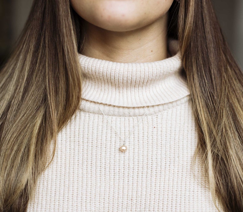 May include: A close-up of a woman wearing a cream-colored turtleneck sweater with a delicate pearl necklace.