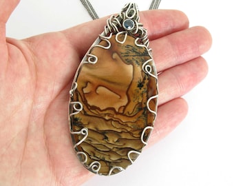 May include: A silver wire wrapped pendant featuring a large, oval-shaped, brown and white agate stone with a blue gemstone accent.