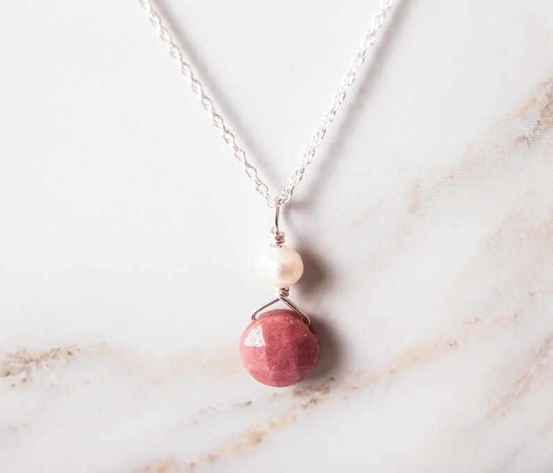 Puede incluir: Un collar de cadena de plata con un colgante de piedra rosa y un acento de perla blanca.