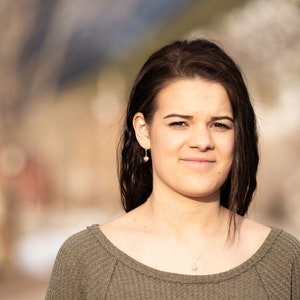 May include: A young woman with long brown hair is wearing a green sweater. She is looking at the camera and smiling slightly. The background is blurred.