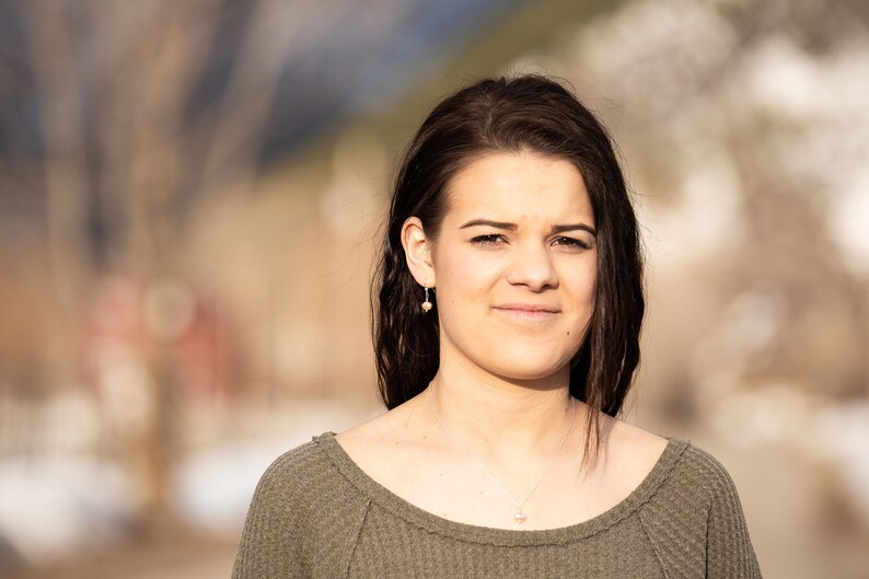 May include: A young woman with long brown hair is wearing a green sweater. She is looking at the camera and smiling slightly. The background is blurred.