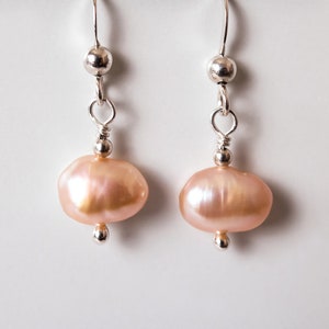 May include: A pair of silver dangle earrings with pink freshwater pearls. The pearls are oval shaped and have a slight iridescent sheen.