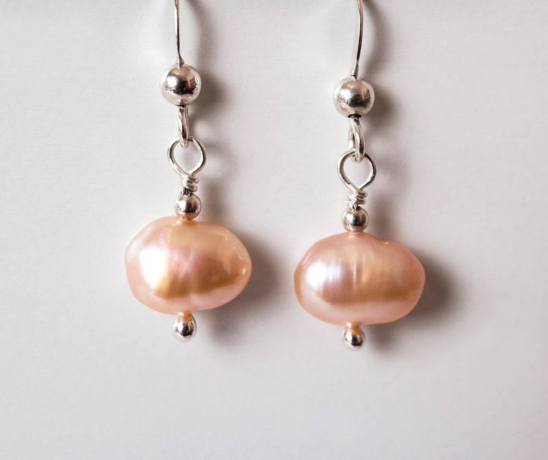May include: A pair of silver dangle earrings with pink freshwater pearls. The pearls are oval shaped and have a slight iridescent sheen.