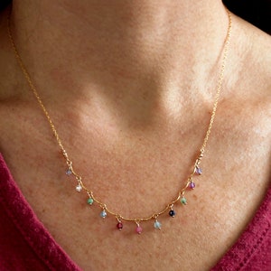 May include: A gold chain necklace with small, colorful gemstones hanging from it. The necklace is worn by a person with a red shirt.