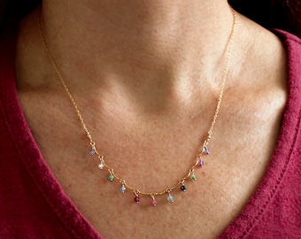 May include: A gold chain necklace with small, colorful gemstones hanging from it. The necklace is worn by a person with a red shirt.
