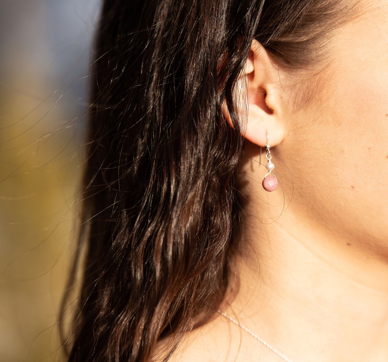 Puede incluir: Un par de pendientes de plata con una piedra preciosa rosa y una peque&ntilde;a perla colgante. Los pendientes los lleva una persona con el pelo largo y casta&ntilde;o.