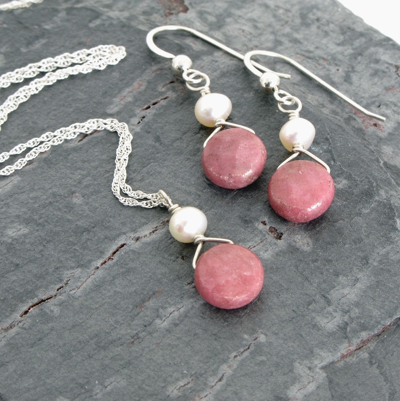 Puede incluir: Un collar y pendientes de plata con piedras rosas y blancas. El collar tiene una cadena delicada y un colgante con una piedra rosa y una perla blanca. Los pendientes tienen un dise&ntilde;o similar con una piedra rosa y una perla blanca.