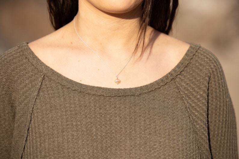 May include: A close-up of a woman wearing a green waffle knit sweater with a delicate silver chain necklace featuring a small pearl pendant.