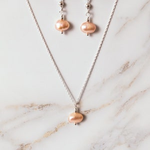 May include: A delicate silver chain necklace with a single blush-colored pearl pendant. Matching earrings with blush-colored pearl drops hang from silver hooks.