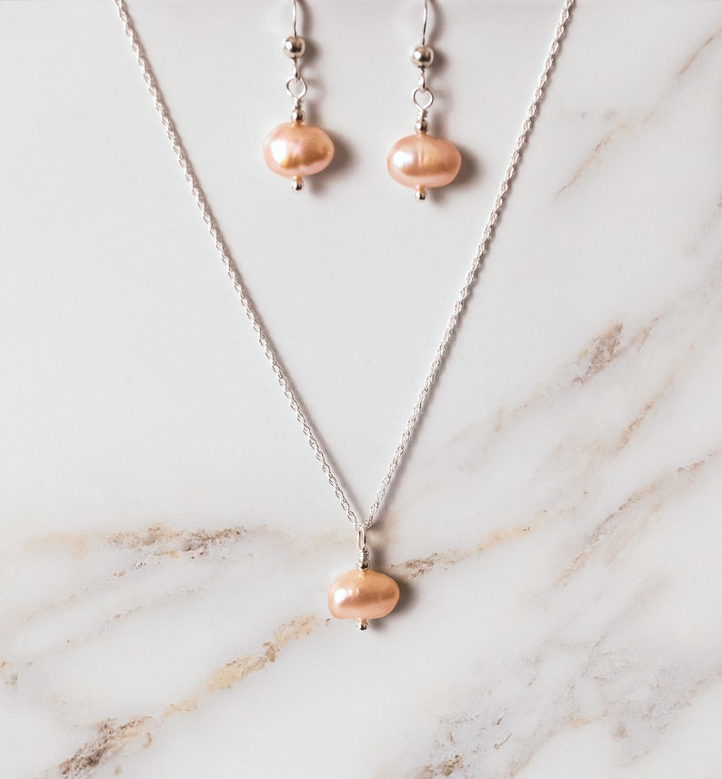 May include: A delicate silver chain necklace with a single blush-colored pearl pendant. Matching earrings with blush-colored pearl drops hang from silver hooks.