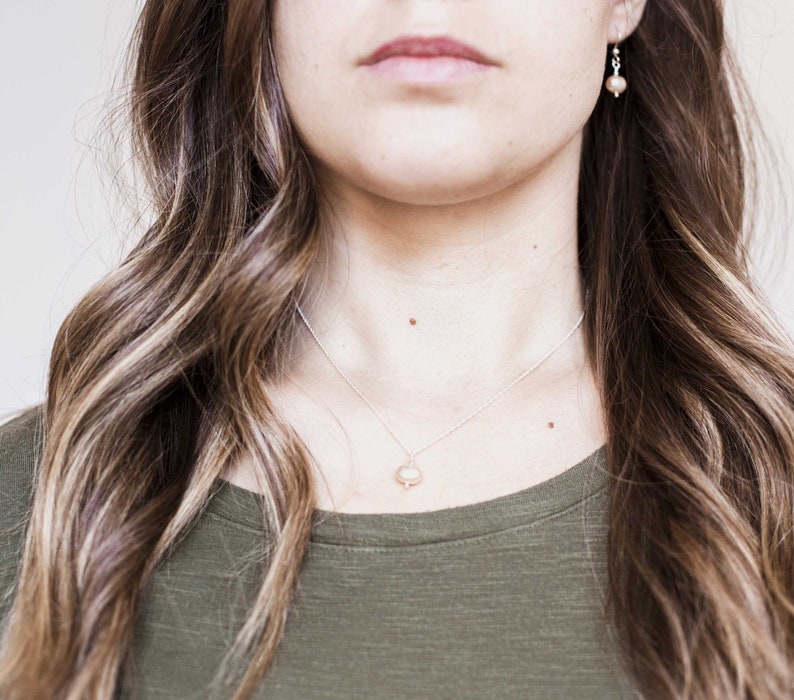 May include: A woman with long brown hair wearing a green shirt and a silver necklace with a pearl pendant. She is also wearing small pearl earrings.
